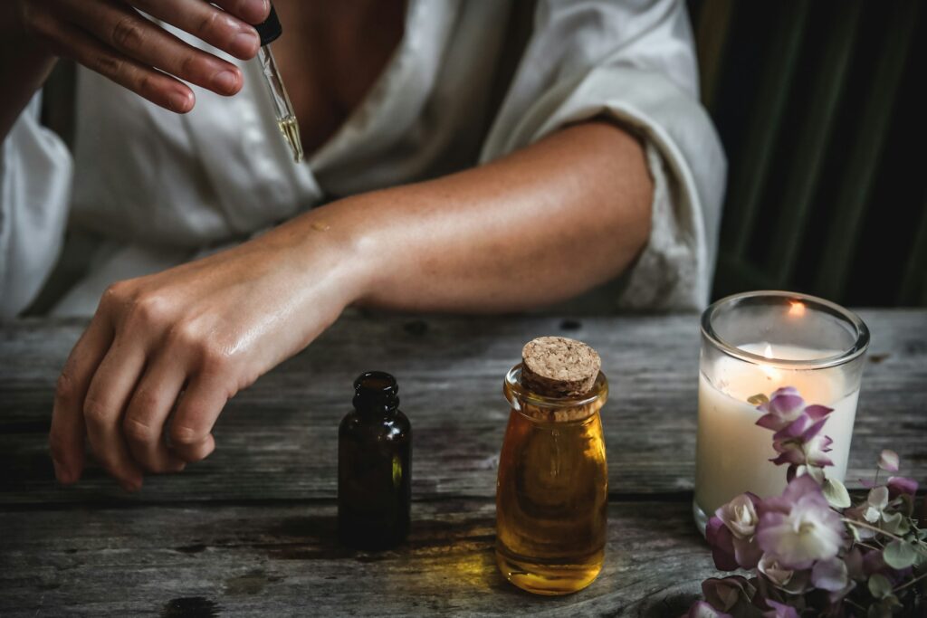 Eine Frau gibt mit einer Pipette ätherisches Öl auf ihren Unterarm; daneben stehen Ölfläschchen, eine brennende Kerze und Blüten – ätherische Öle beinhalten Pflanzen- und Ölwesen.