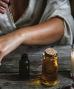 Eine Frau gibt mit einer Pipette ätherisches Öl auf ihren Unterarm; daneben stehen Ölfläschchen, eine brennende Kerze und Blüten – ätherische Öle beinhalten Pflanzen- und Ölwesen.